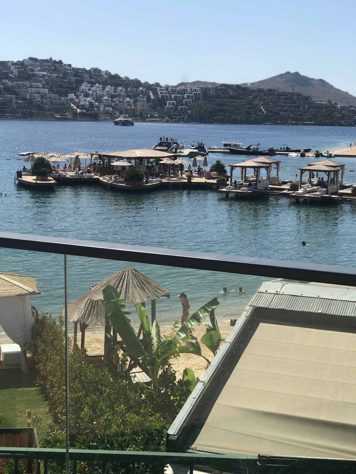 A serene water view features boats docked at a marina, with sunbathers enjoying the beach. Surrounding greenery and umbrellas create a relaxing atmosphere. In the background, hillside villas are visible against a clear blue sky, enhancing the tranquil scene.