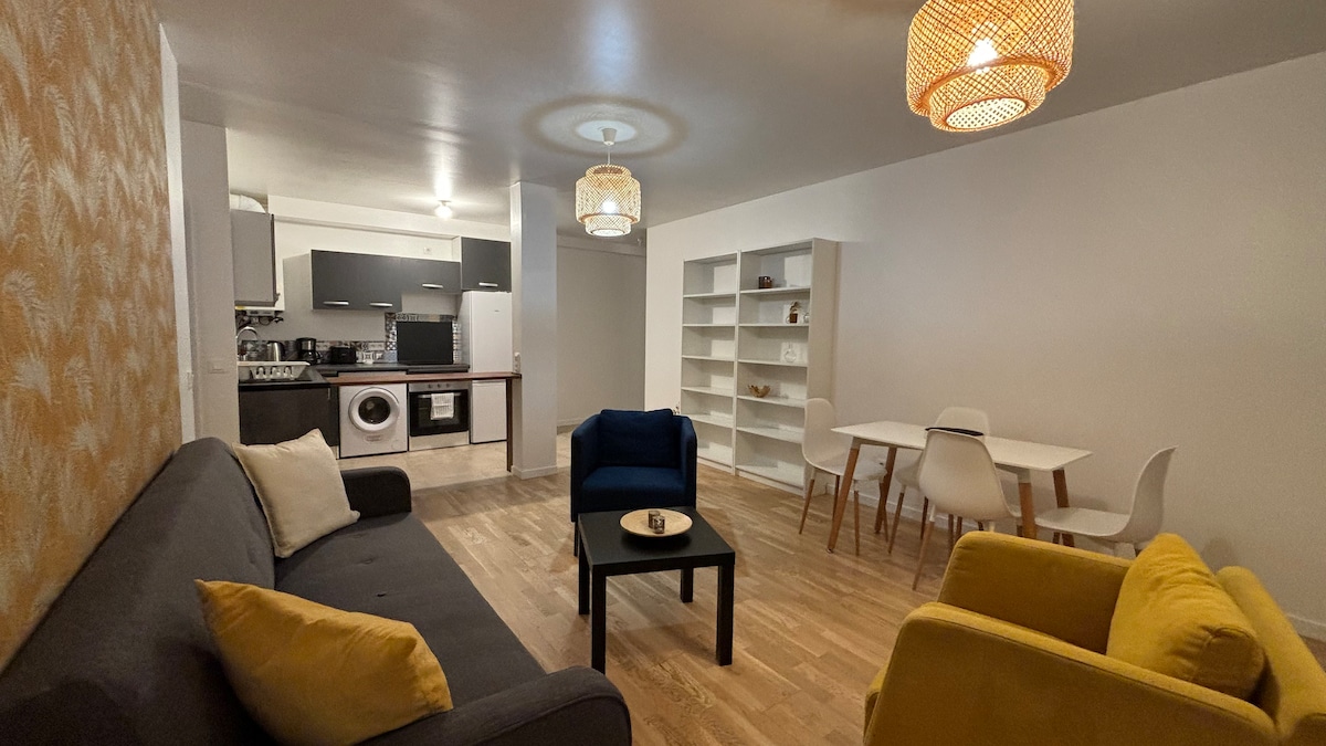 A welcoming living area features a dark grey sofa alongside a blue armchair and a bright yellow armchair. A circular black table sits in the center, with a decorative item placed atop. Soft lighting is provided by hanging fixtures, and a white shelving unit line one wall.