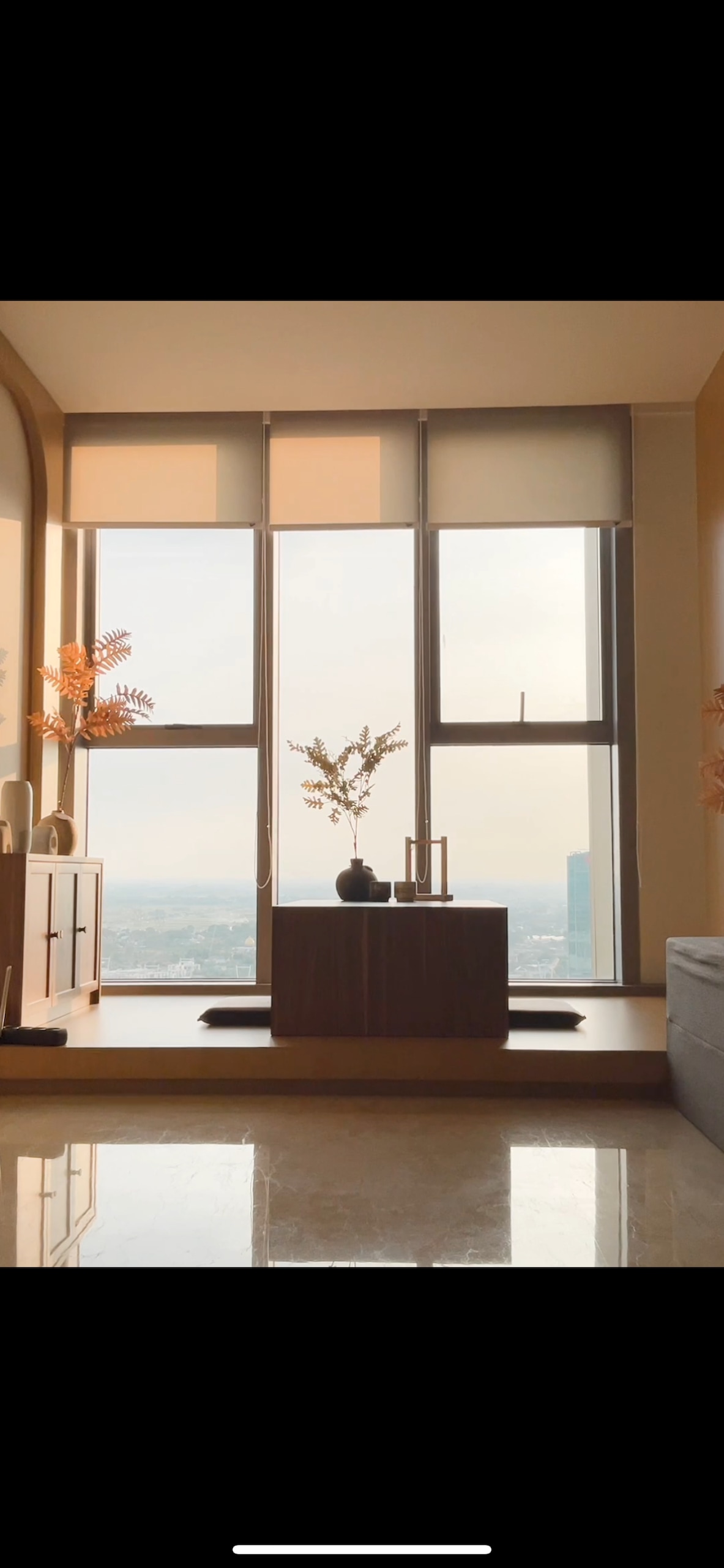 A calming living area features a low wooden table accompanied by a decorative vase and minimalistic accessories. Large windows provide ample natural light and offer a view of the city skyline. Bright, neutral tones create a serene atmosphere.