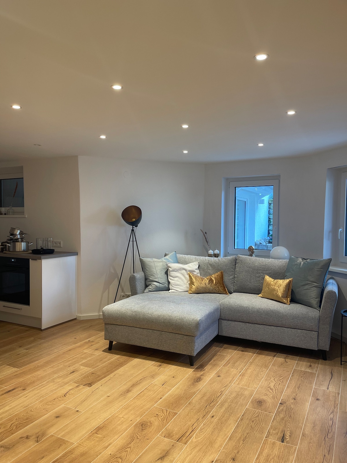 A light and airy living area features a spacious gray sectional sofa adorned with decorative cushions. A sleek black floor lamp stands beside the sofa. Soft lighting from recessed ceiling fixtures enhances the warm wooden floor, while a window offers an outdoor view.