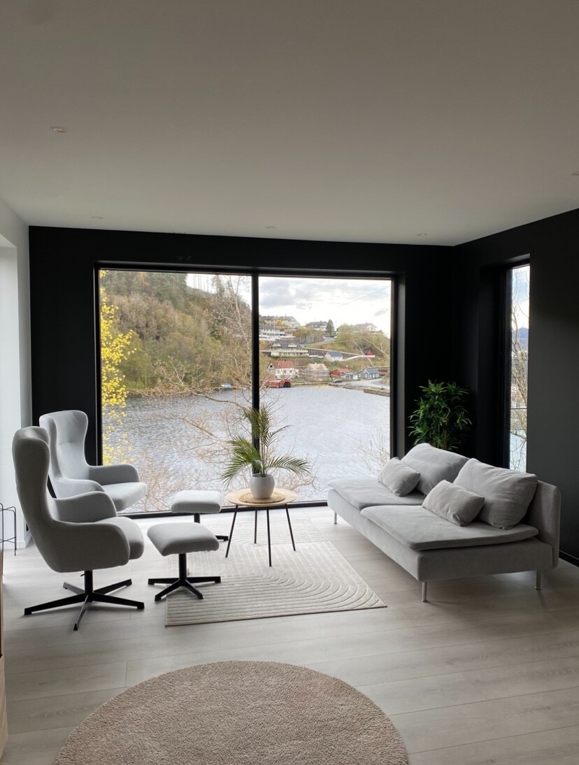 A modern living area features a large window offering a scenic view of the water and surrounding greenery. A gray sofa and a stylish armchair are arranged around a small round table, while a light-toned area rug adds warmth to the space.