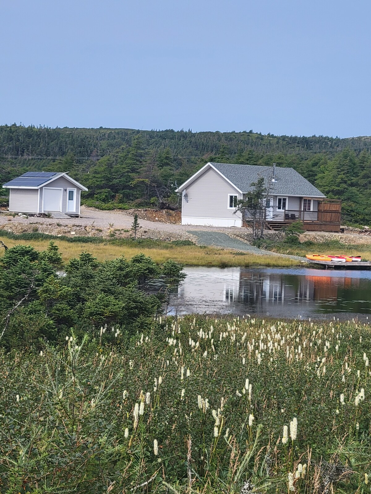 A serene landscape showcases a cabin and an additional outbuilding near the water's edge. Lush greenery surrounds the area, and kayaks rest beside the calm lake, reflecting the clear sky. The natural setting invites relaxation and exploration in a peaceful environment.