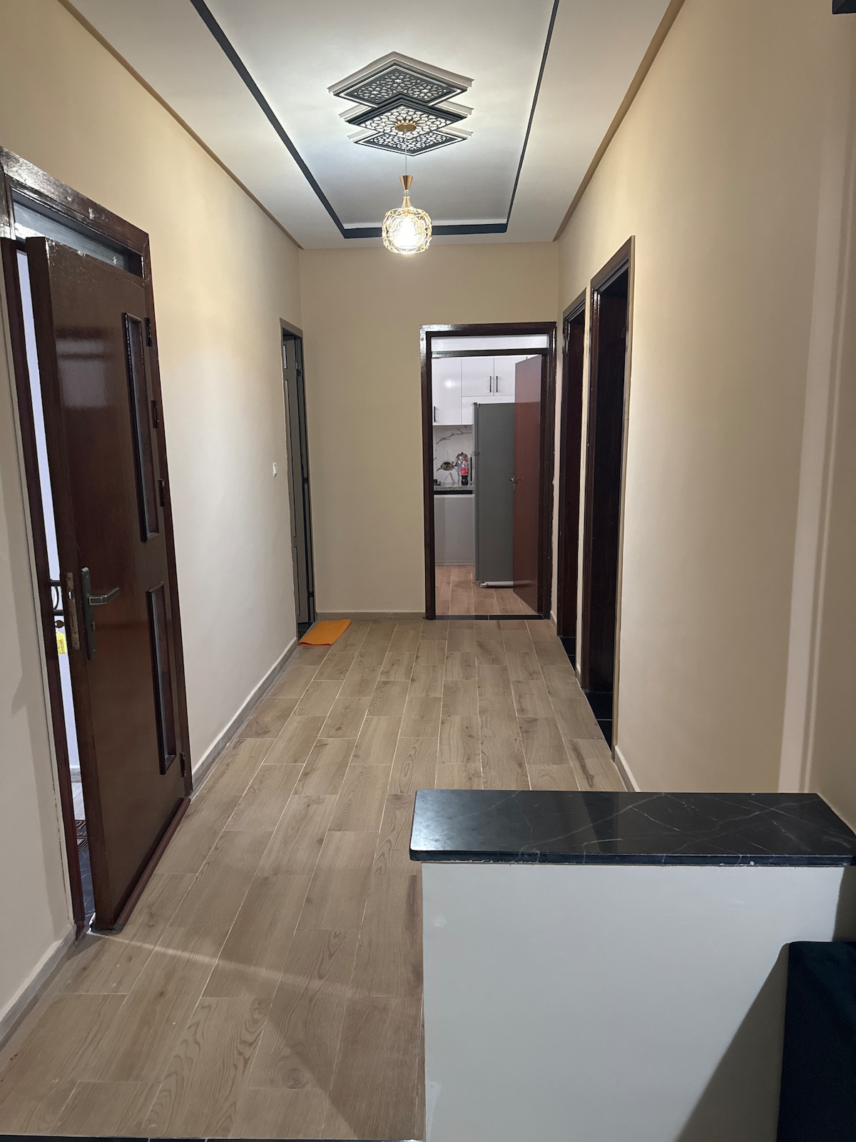 A well-lit hallway displays wooden flooring and neutral wall colors. Multiple doors line either side, leading into various rooms. A glimpse of the kitchen is visible at the end of the corridor, where modern cabinetry and appliances can be seen.