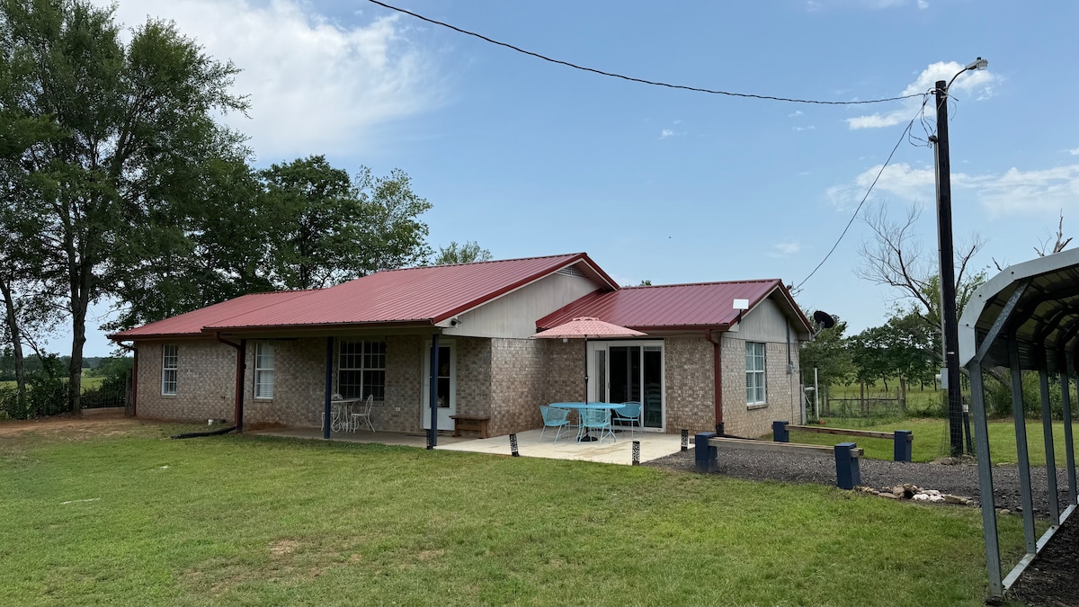 The exterior of the ranch-style home is shown, featuring a red metal roof and a brick facade. A spacious patio area with outdoor seating is visible, alongside well-maintained green grass. Surrounding trees and open fields contribute to the peaceful countryside setting.