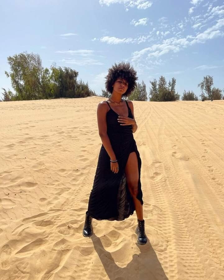 A figure is seen standing confidently on a sandy dune under a clear blue sky. The flowing black dress features a thigh-high slit and is complemented by black boots. Sparse vegetation and distant trees are visible in the background, enhancing the scene's natural setting.