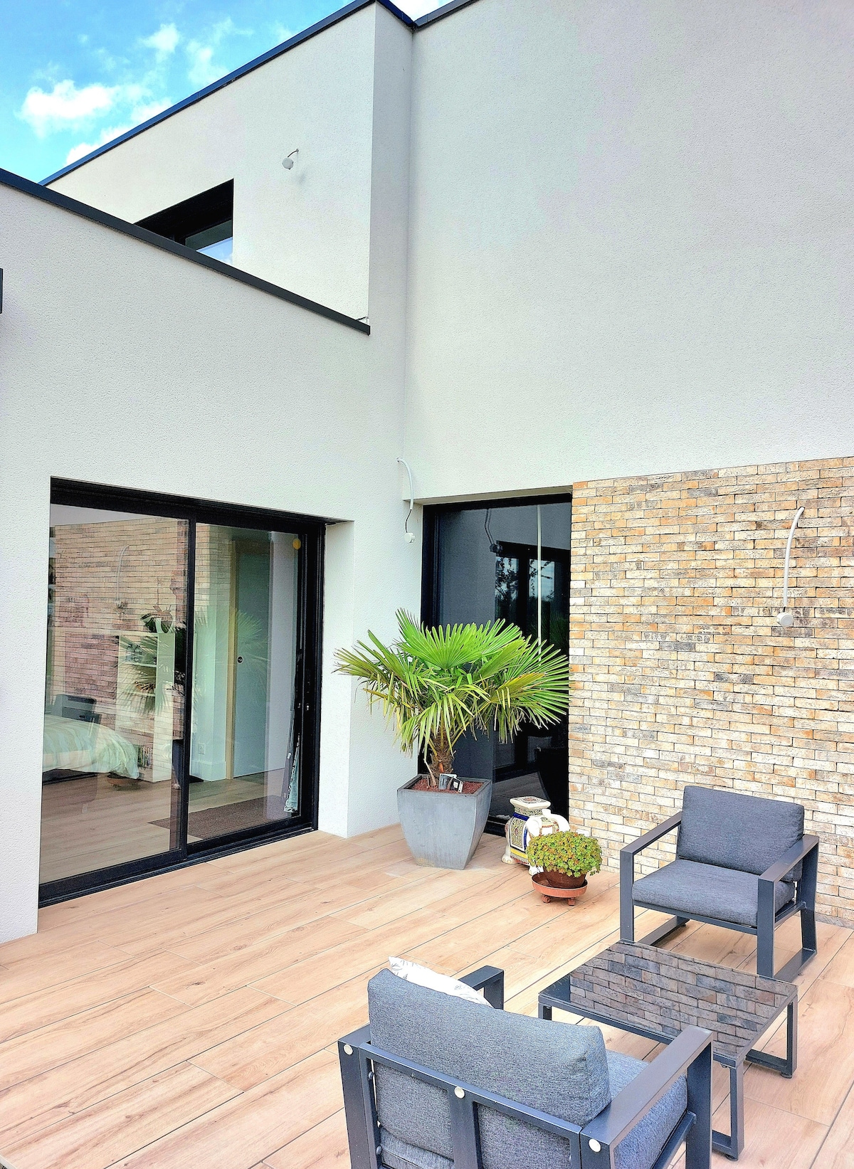 A serene terrace is presented, featuring a pair of black lounge chairs and a small table. A potted palm adds a touch of greenery. Glass doors lead into the interior space, revealing a light-filled room with stone accents and wooden flooring.