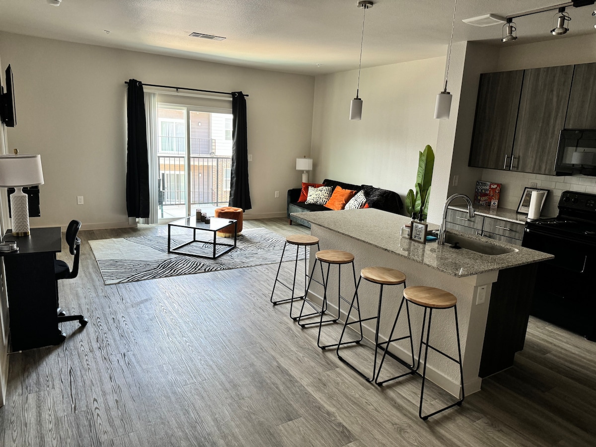 An open living space features a modern kitchen with a breakfast bar and four wooden stools. A comfortable sofa is adorned with orange and black pillows, while a large sliding door allows natural light and access to a balcony. A neutral area rug adds texture to the flooring.
