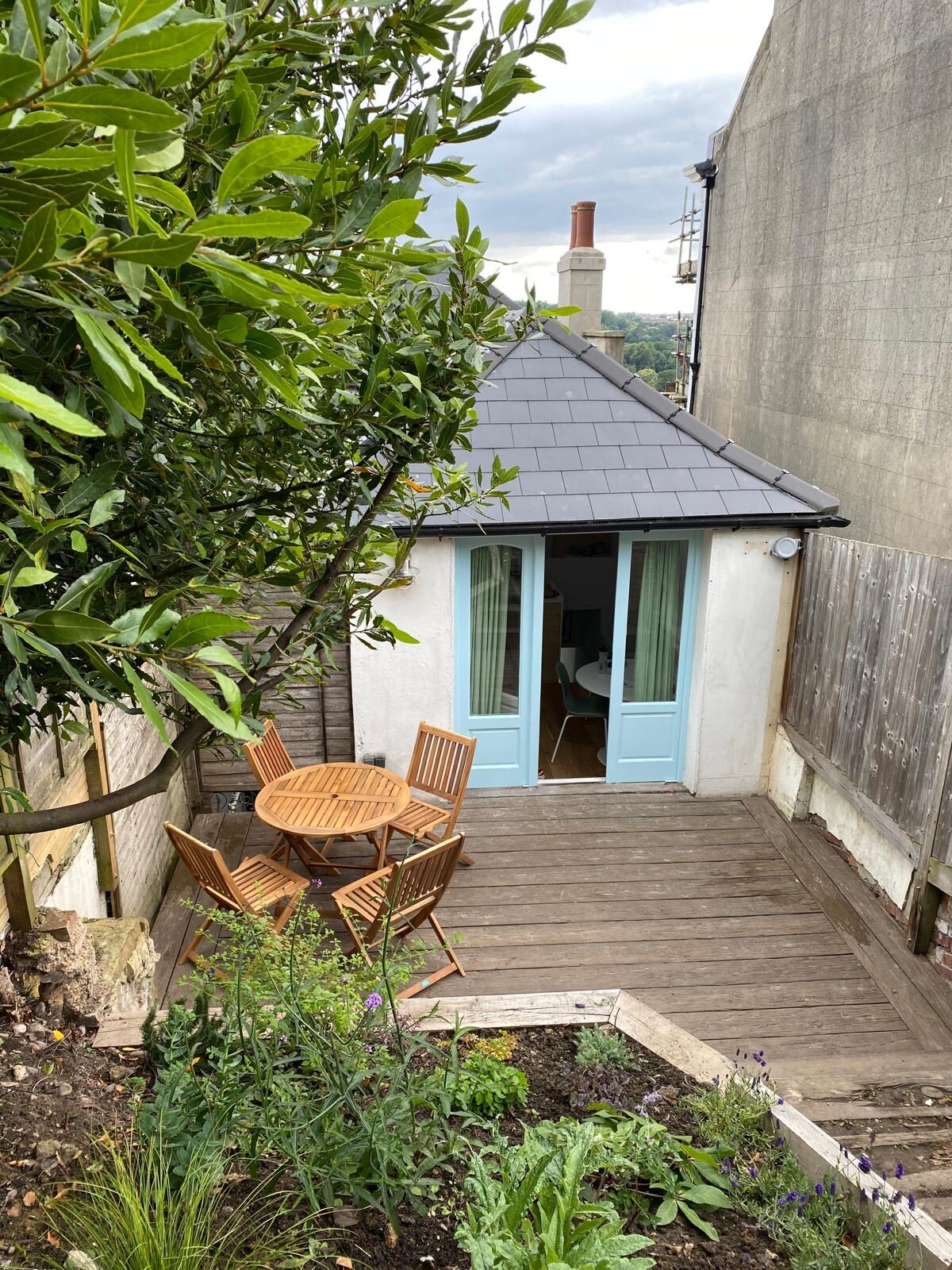 A quaint outdoor seating area is shown, featuring a small round table surrounded by four wooden chairs. French doors lead into the interior, partially obscured by greenery. The deck is nestled among plants, adding a touch of nature to the peaceful space.