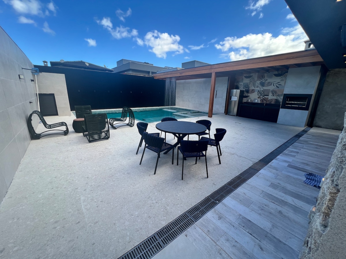 An outdoor leisure area features a pristine swimming pool surrounded by modern seating arrangements. A circular dining table is positioned under a covered area, while two loungers offer relaxation options nearby. Bright blue skies enhance the inviting atmosphere of the space.