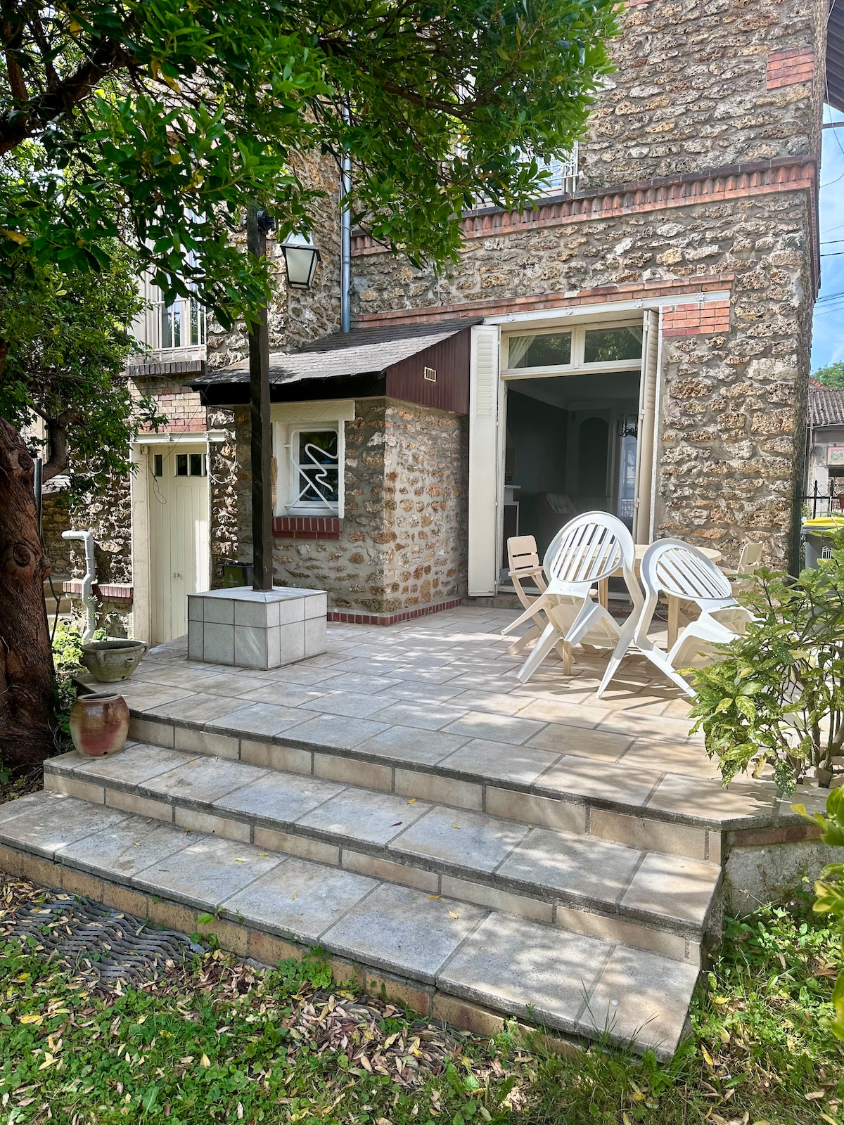 A patio area is featured, showcasing stone steps leading to a spacious outdoor setting. A small table with four white chairs is arranged on the terrace, framed by lush greenery. The stone facade of the house is visible in the background, emphasizing its architectural character.