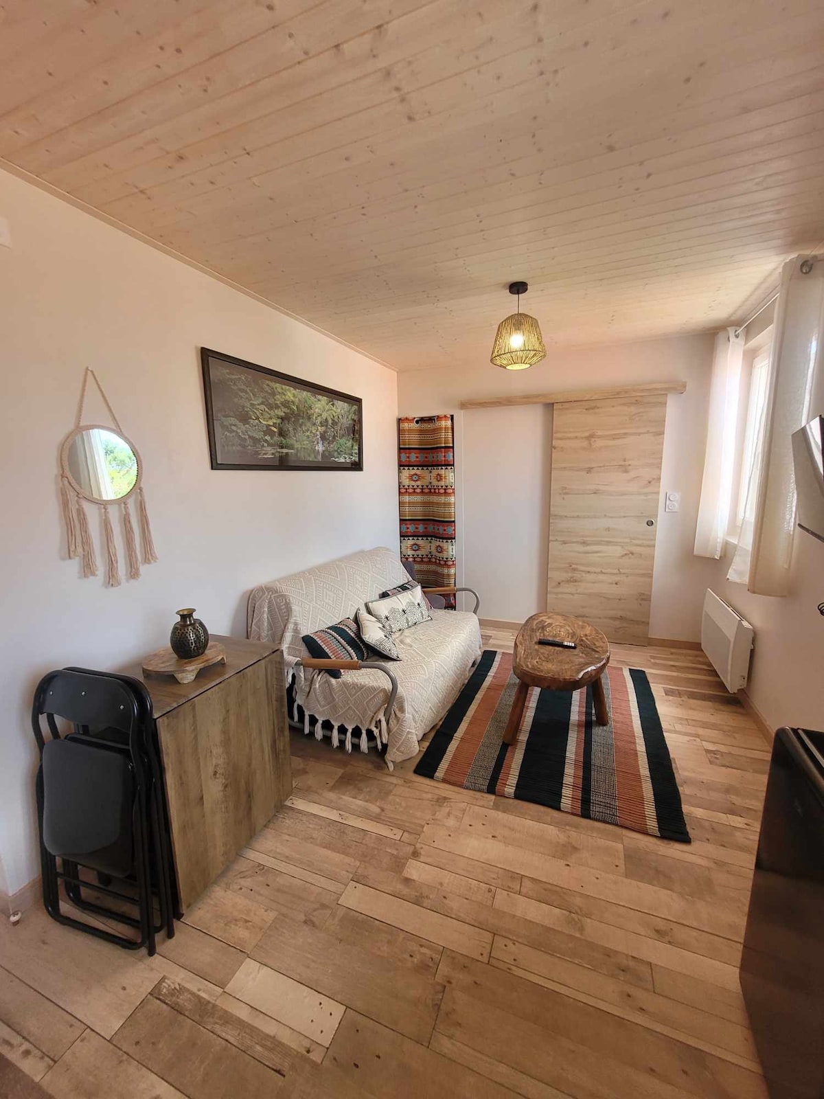 A inviting living area is shown, featuring a comfortable sofa draped with a light throw. A round coffee table sits on a patterned rug, with wooden flooring providing a warm tone. Natural light enters through the windows, which are adorned with sheer curtains.