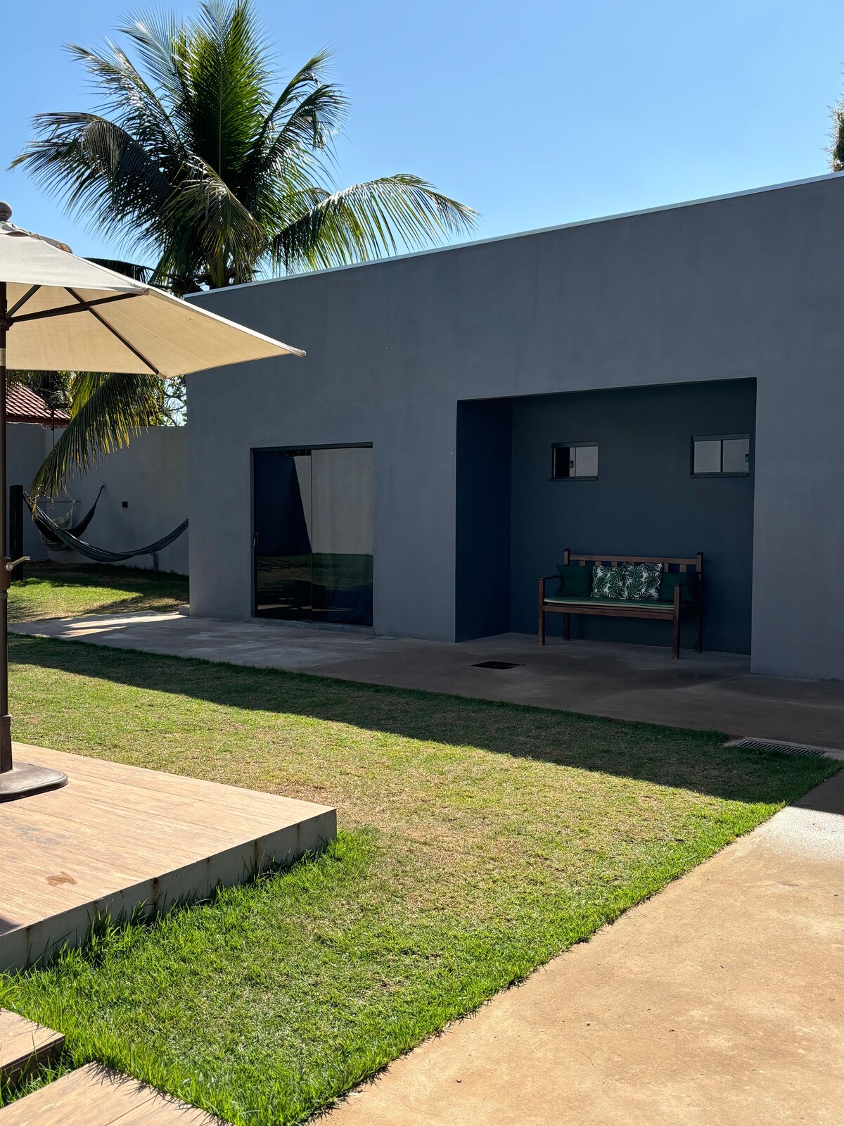 A shaded outdoor area features a lounge chair and a hammock, framed by swaying palm trees. The gray exterior of the house contrasts with the lush green grass. An entrance with a glass door and a window allows natural light to enhance the area.