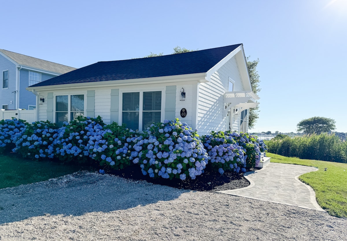 A charming cottage exterior is framed by vibrant blue hydrangea bushes, adding color to the landscape. The building features a white clapboard facade with multiple windows. A paved pathway leads to the entrance, surrounded by lush greenery and a clear sky.