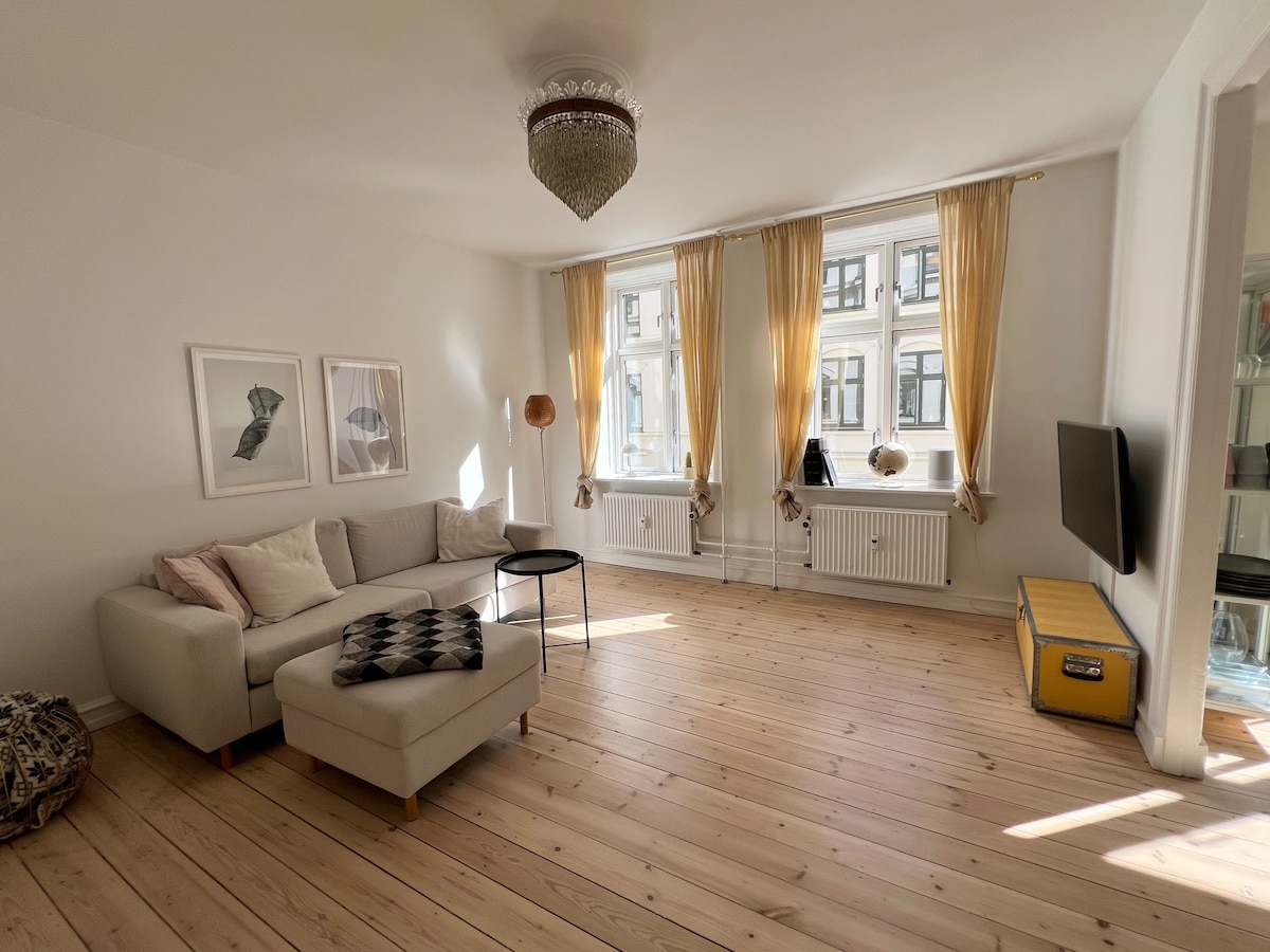A spacious living area features a light-colored sofa with a patterned throw. Two large windows with sheer curtains allow natural light to fill the room. A modern black side table sits beside the sofa, and a flat-screen TV is positioned on a nearby wall.