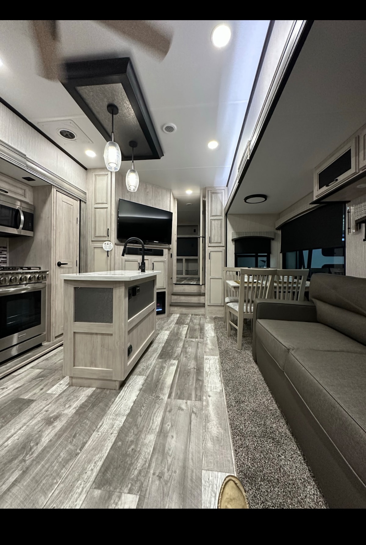A modern tiny home interior is displayed, featuring an open layout with light wood cabinetry and a spacious island. The living area includes a comfortable sofa and a dining table, with large windows allowing natural light to fill the room, enhancing the inviting ambiance.