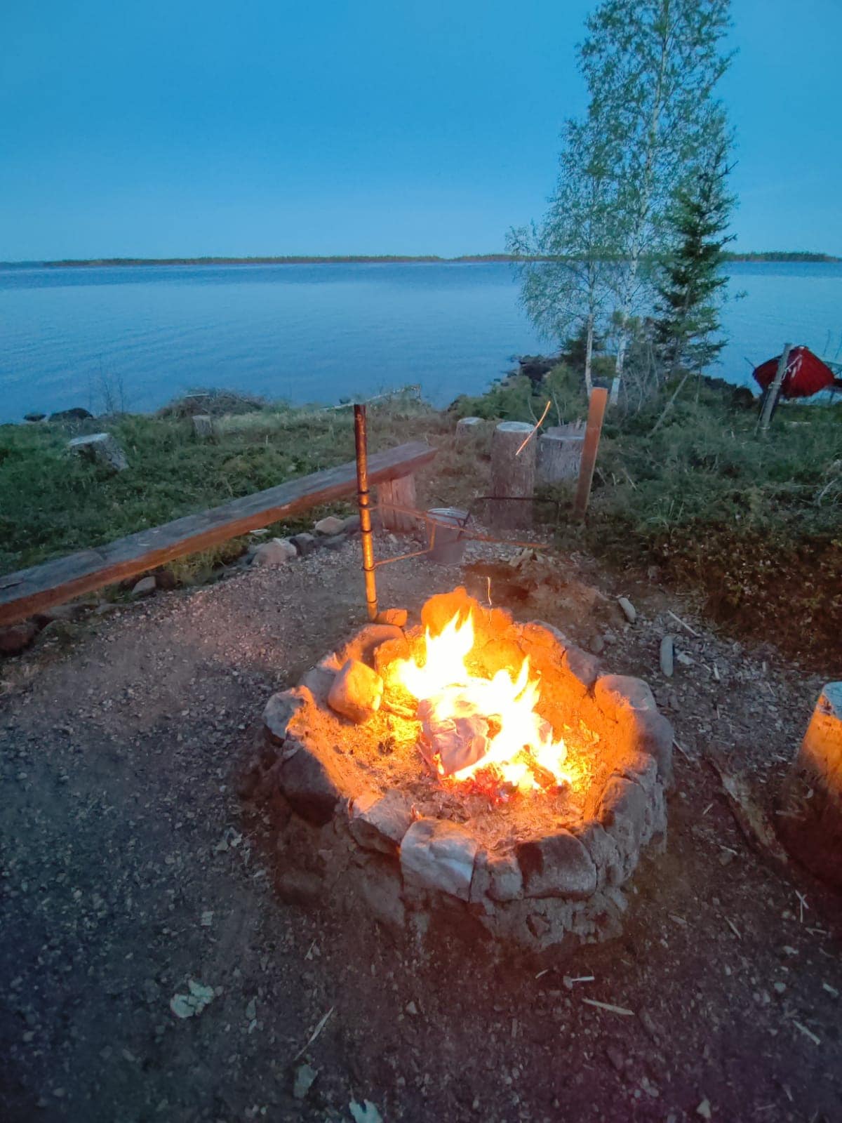 A fire pit, constructed from stones, emits a warm glow as flames dance above the wood. The setting features a serene lake in the background, surrounded by greenery, with a hint of a tent visible in the distance under the evening sky.