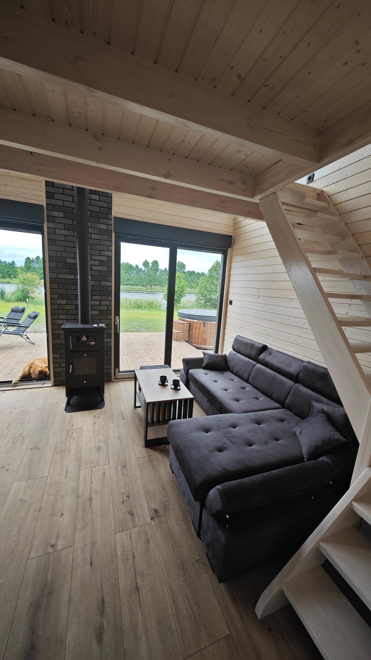 A bright interior is observed with a gray sectional sofa positioned in front of a coffee table. Large glass doors allow natural light and views of the outdoor scenery, while a small wood stove provides warmth. A staircase leads to the upper level.