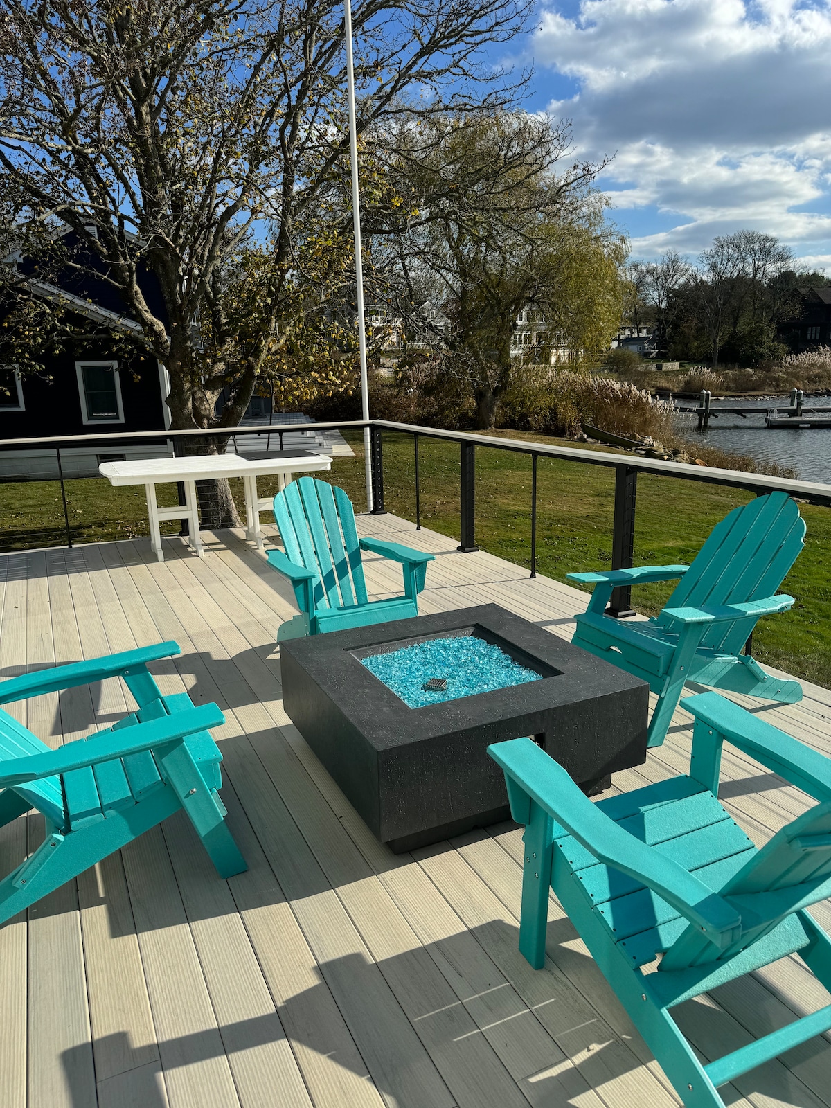An outdoor deck features a square propane fire pit surrounded by four turquoise Adirondack chairs. A white table is positioned in the background, with a grassy area and a pond visible beyond the railing. Bright clouds can be seen against the blue sky.
