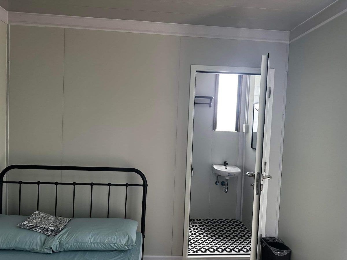 A simple room features a metal bed with a light blue mattress. A folded blanket is placed on the bed. A mirror and a small sink are visible in the adjoining bathroom, with a door leading to the bathroom being partially open.