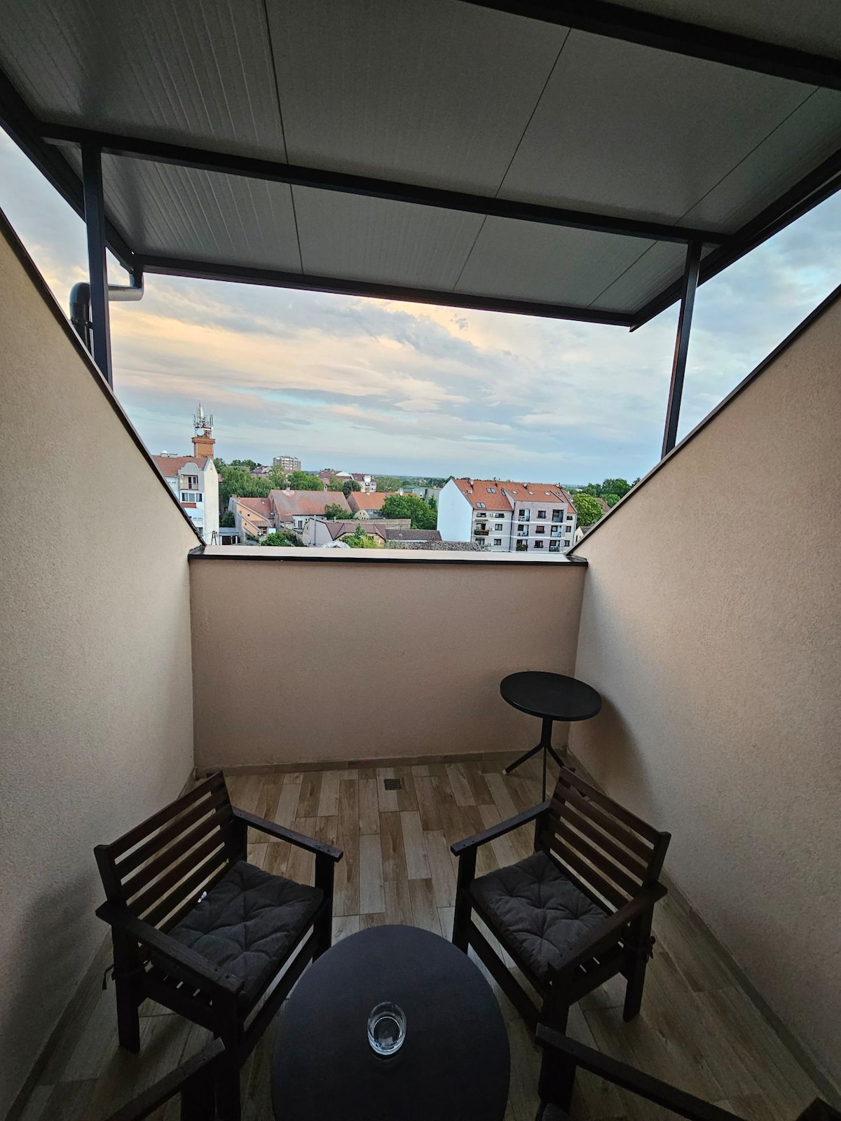A small balcony features two wooden chairs and a round table. The view showcases a neighborhood with colorful rooftops against a cloudy sky. The flooring consists of light wood planks, creating a serene outdoor space for relaxation.