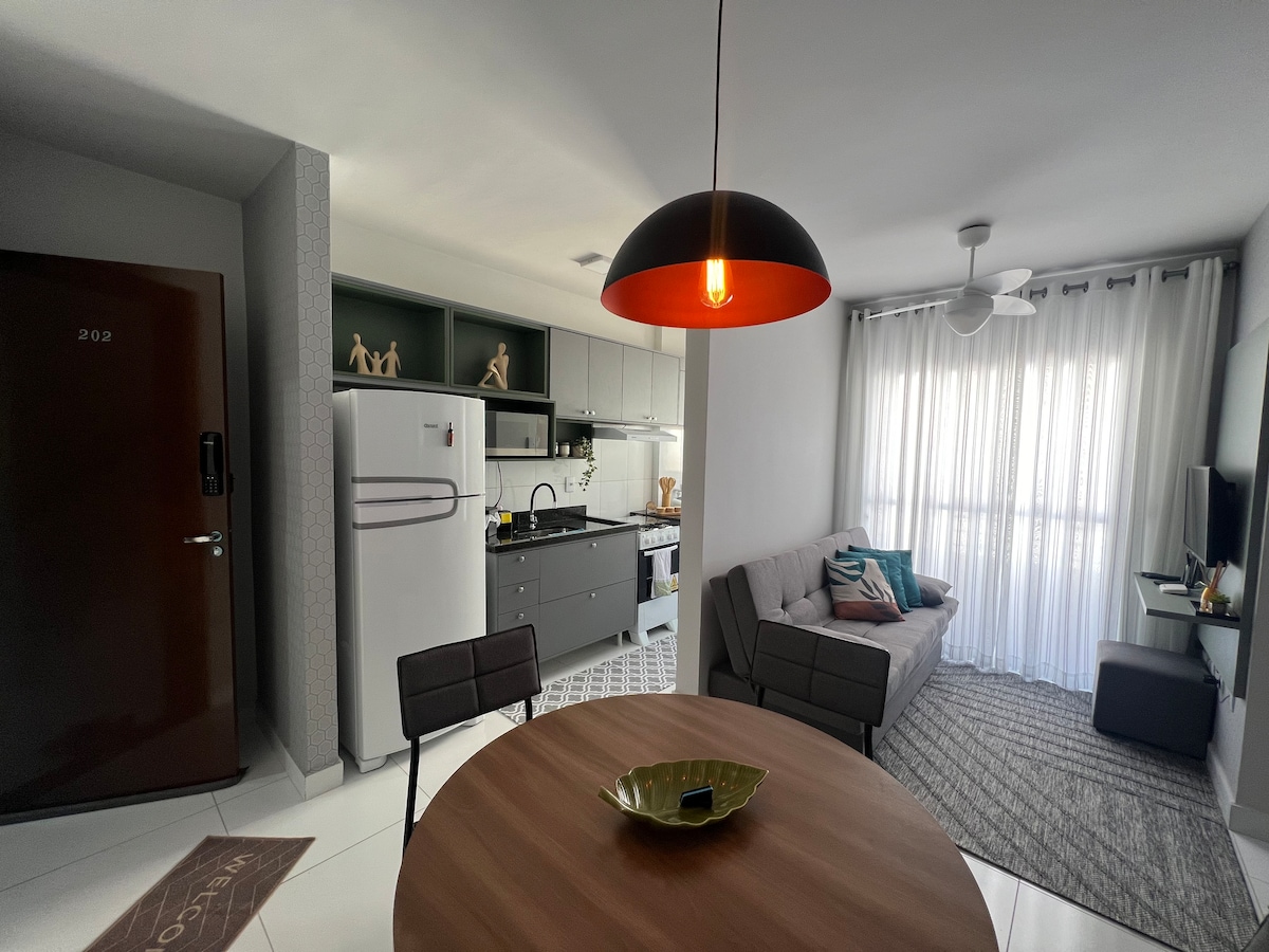 An open-plan living space is presented, featuring a circular dining table with dark chairs. A modern kitchen is visible, equipped with a white refrigerator and black countertops. Natural light filters through sheer curtains onto a cozy sofa and a small TV stand with a television.