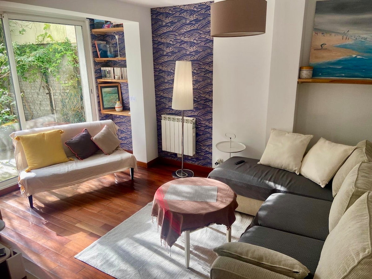 A cozy living area showcases a sectional sofa and a small coffee table placed on a soft rug. Natural light enters through glass doors leading to the garden outside. A lamp and a bookshelf are visible, enhancing the warm ambiance.