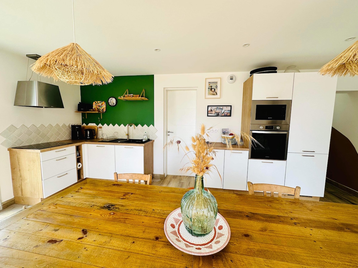A well-equipped kitchen features light wood cabinetry and modern appliances, including an oven and coffee maker. A large wooden dining table with chairs is centered under woven pendant lights. Decorative plants and wall art add a touch of style to the room.