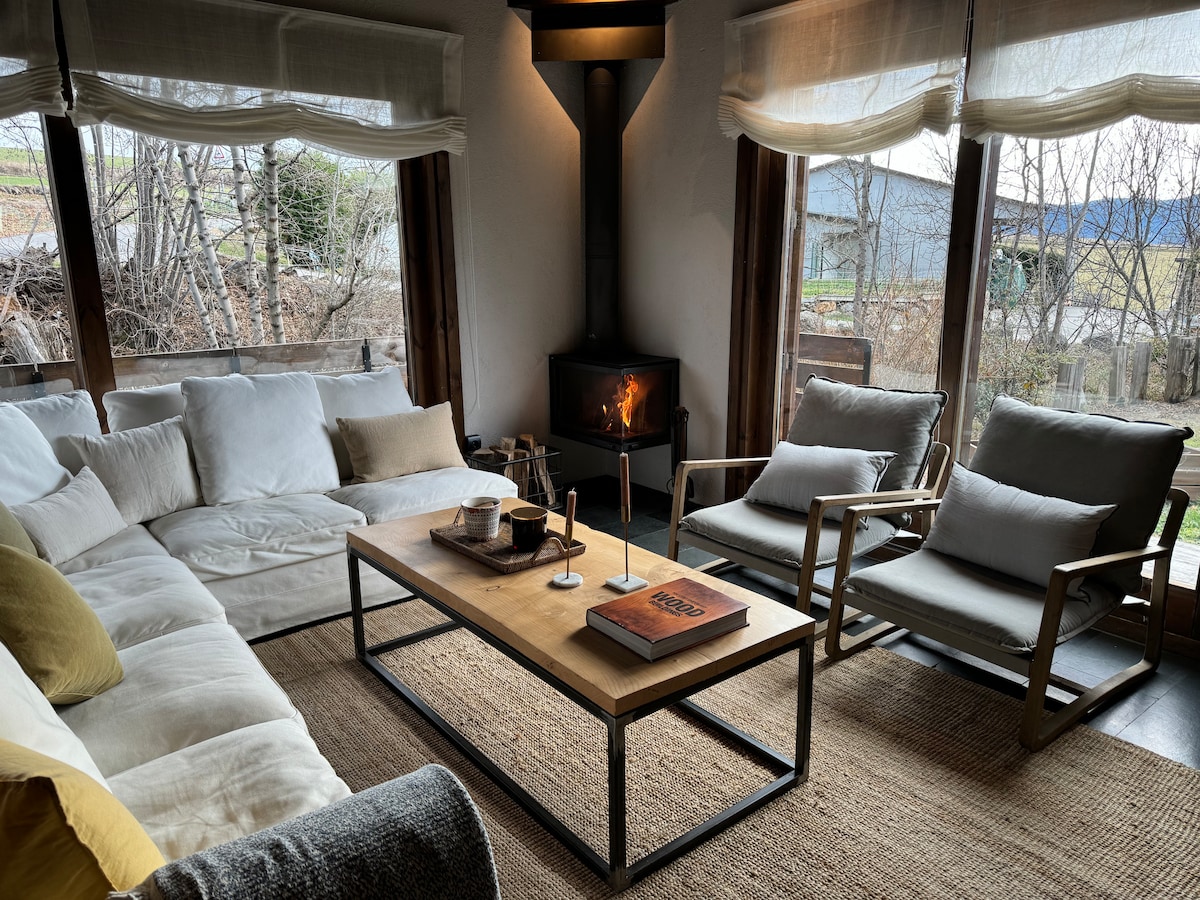 A cozy living area features a large white sectional sofa and two armchairs placed around a wooden coffee table. A fireplace with a gentle flame provides warmth. Large windows offer views of the surrounding landscape, and natural light fills the space.