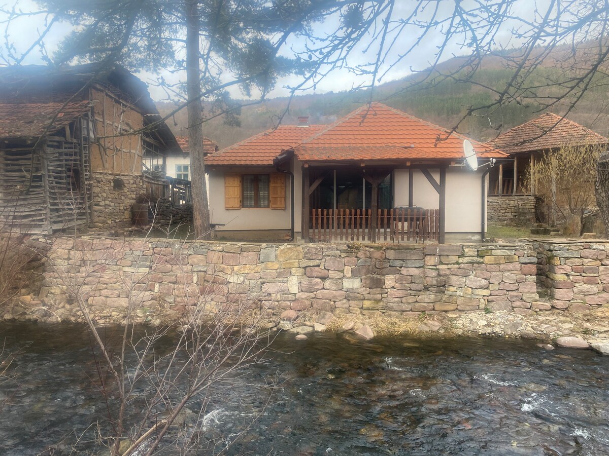 A charming house is positioned beside a gently flowing river, showcasing a warm wooden porch with a tiled roof. Natural stone walls and traditional architecture are complemented by surrounding trees, creating a serene setting in a picturesque landscape.
