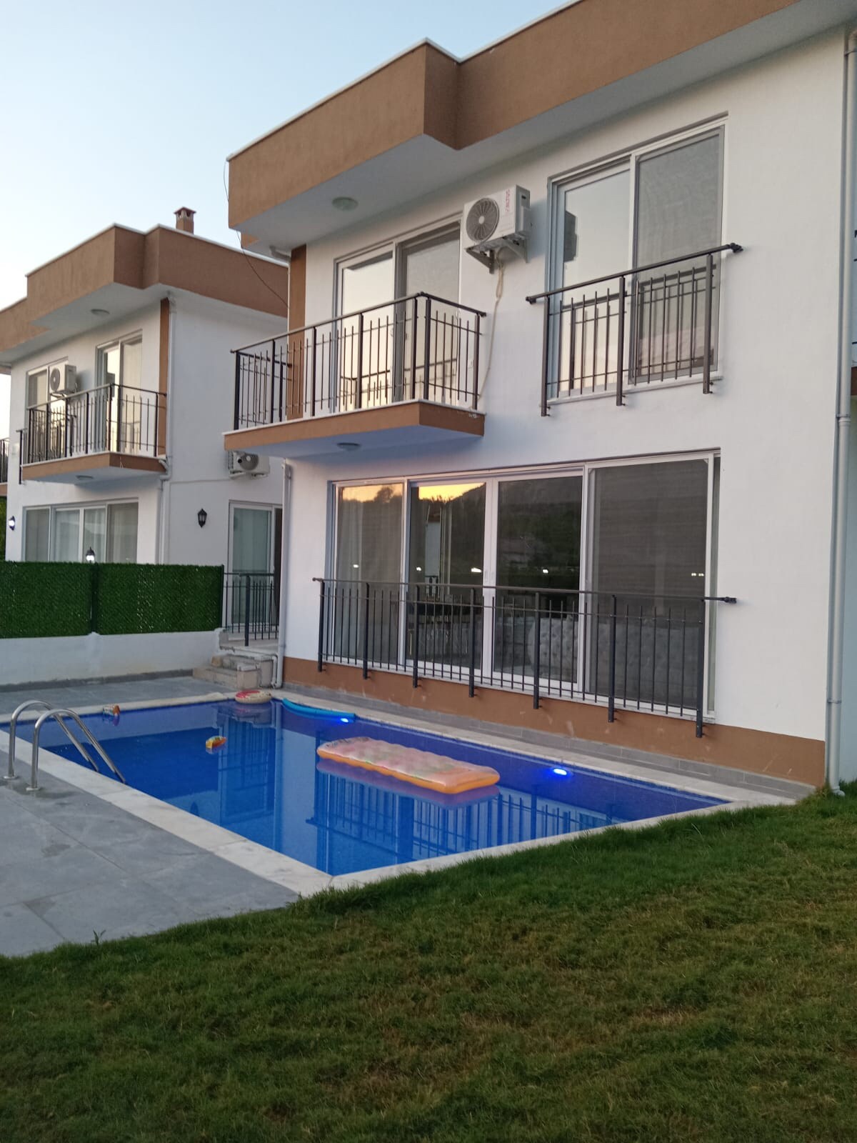 A modern villa exterior showcases a private swimming pool with illuminated features. Balconies with black railings are attached to the upper levels, and large glass doors provide a view into the living spaces. Lush grass borders the pool area, offering a relaxed outdoor setting.