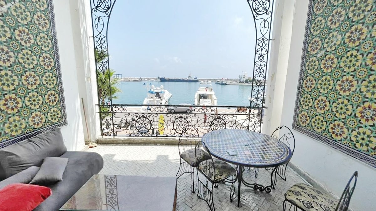 A serene outdoor seating area overlooks a marina, featuring a decorative table surrounded by chairs. Colorful tiles adorn the walls, enhancing the vibrant setting. The view includes boats at dock and the water, inviting a relaxed atmosphere for enjoying the surroundings.