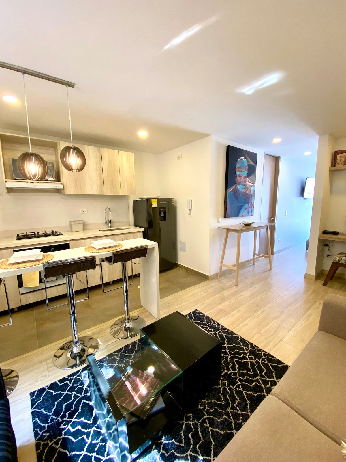 A modern kitchen area is highlighted by sleek cabinetry and a gas stove. Two bar stools are positioned at a stylish peninsula, with a large refrigerator visible nearby. A living space, featuring a neutral sofa and an abstract area rug, offers a welcoming seating arrangement.