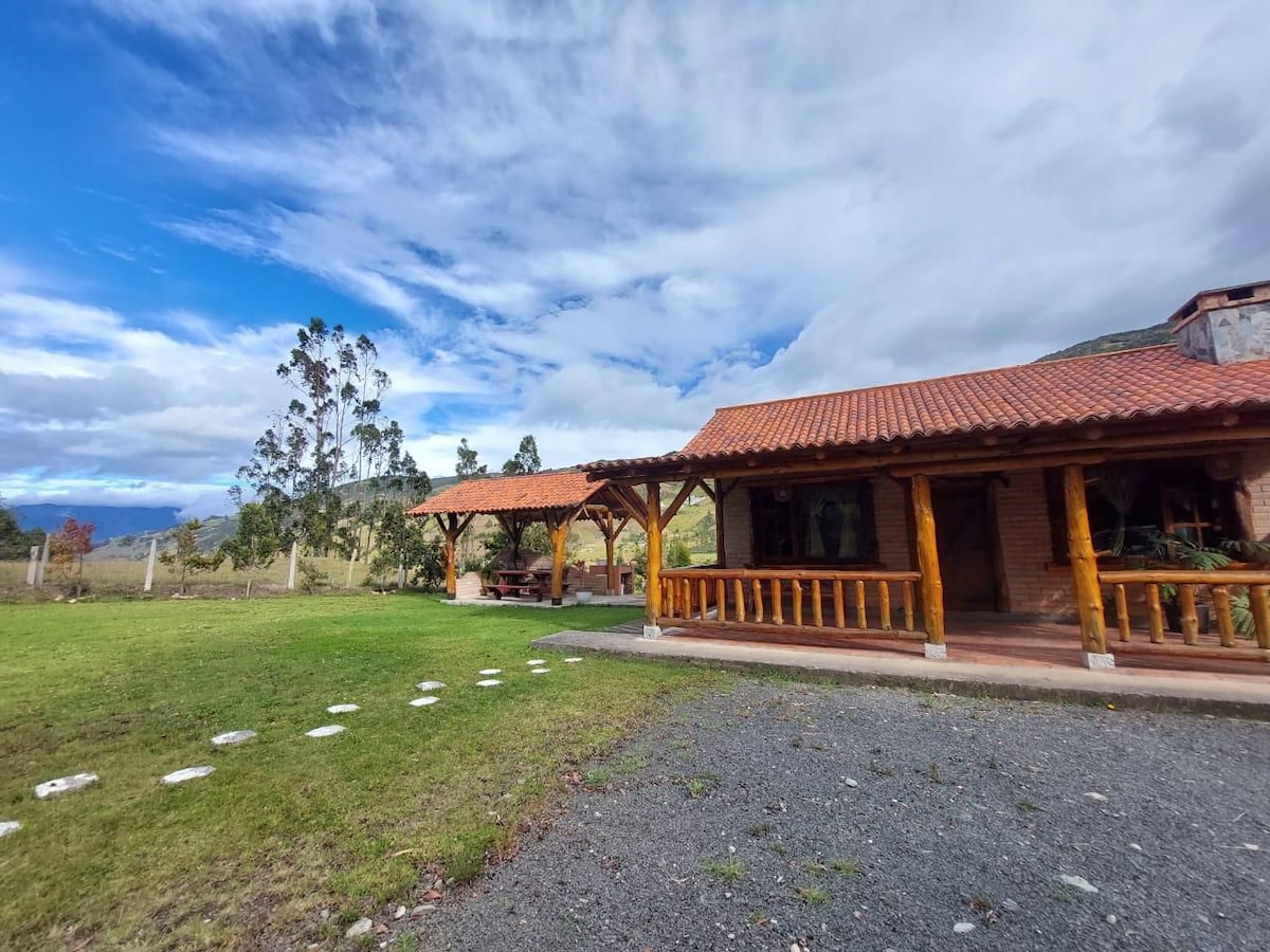 A charming wooden cabin is framed by lush greenery and a blue sky dotted with clouds. Paved pathways lead to the entrance, accompanied by a well-manicured lawn. The structure features a tiled roof and inviting porch areas, perfect for relaxing outdoors.