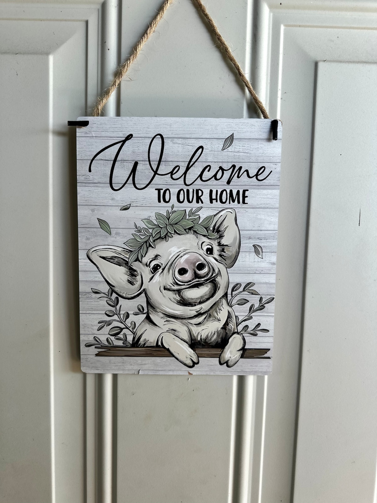 A charming wall sign is hung, displaying a friendly pig illustration surrounded by greenery. The text 'Welcome to Our Home' is scripted above the pig, creating an inviting focal point. The sign is mounted on a plain surface, adding a homey touch.