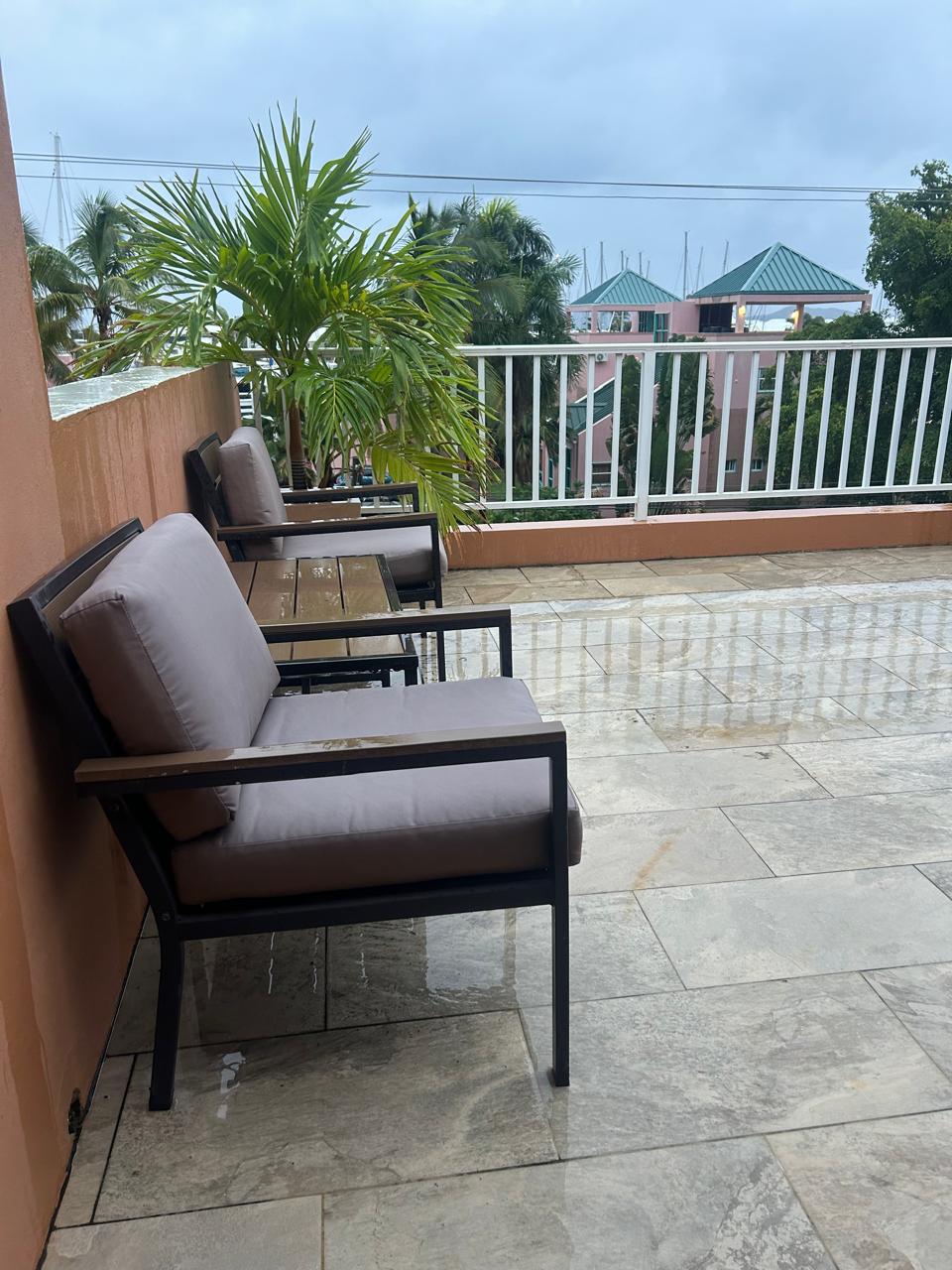 An outdoor patio showcases three minimalist chairs with comfortable cushions, arranged on a tiled surface. A lush palm tree adds greenery, while a railing provides safety and scenic views of the surroundings. The sky appears overcast, suggesting a calm atmosphere.