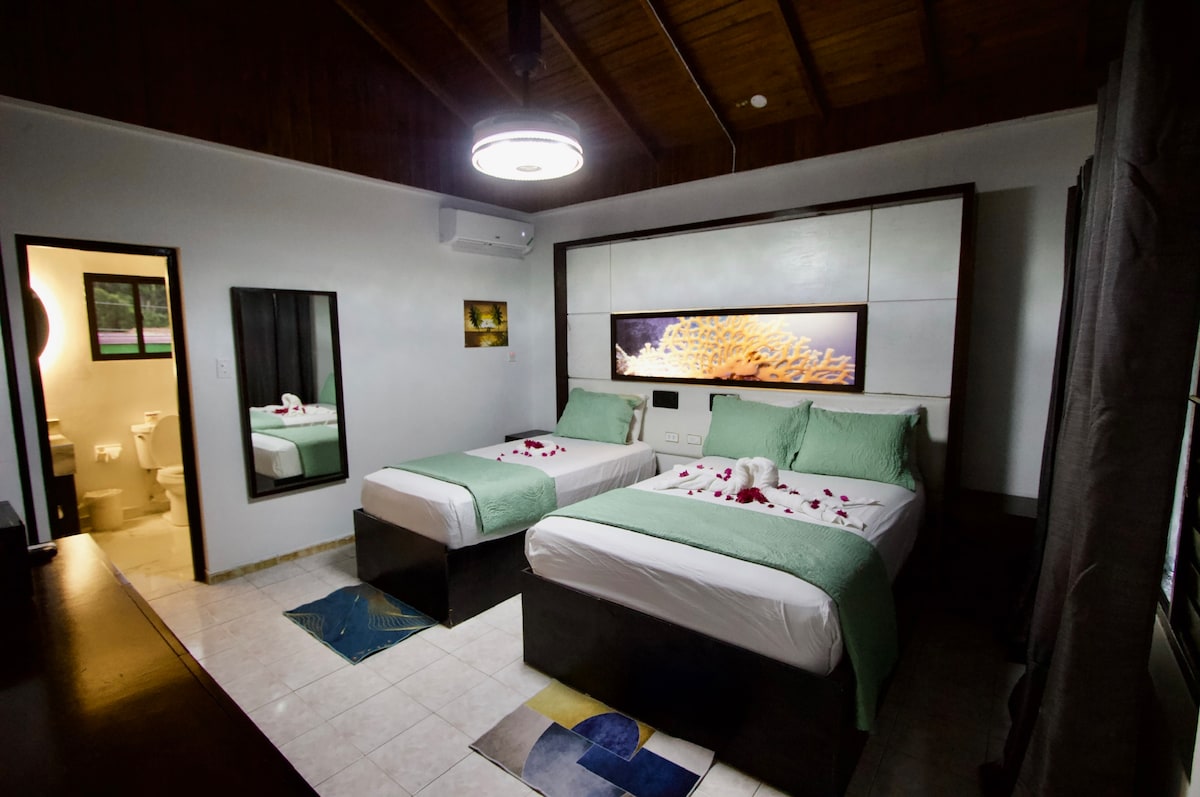 The room features two spacious beds dressed in crisp white linens, with green accents at the foot. A large mirror reflects the room's soft lighting, while a framed artwork adorns the wall. Light streams through a window, creating a relaxed ambiance.