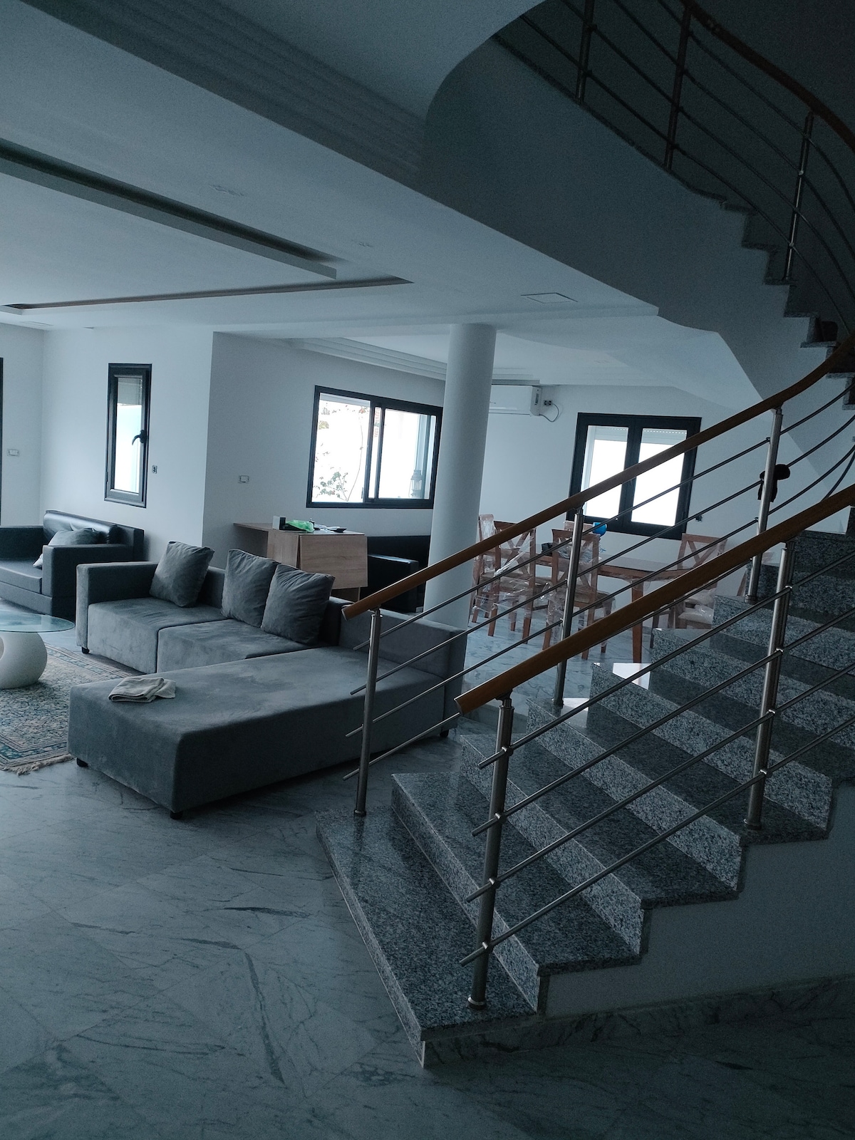 A spacious living area showcases a soft gray sectional sofa opposite a wooden dining table, which is surrounded by several chairs. Natural light enters through multiple windows, highlighting the marble flooring. A staircase with a metal railing leads to the upper level.