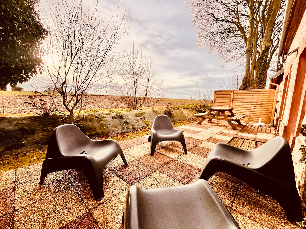 The outdoor terrace features modern seating arranged on a textured surface, providing a space for relaxation. A picnic table is positioned in the background, surrounded by bare trees and natural greenery. The open view captures the expansive landscape beyond.