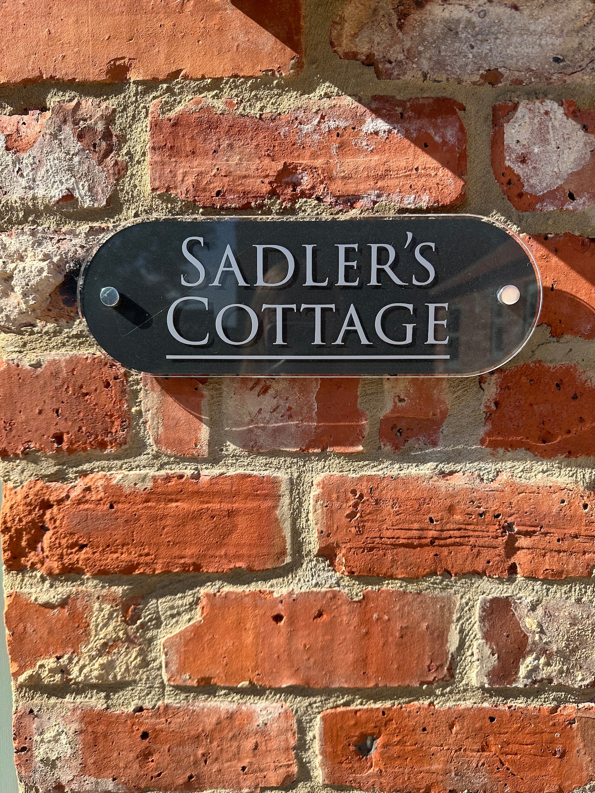A clear sign reading 'Sadler's Cottage' is mounted on a textured brick wall, showcasing the warm tones of the surrounding bricks. The sign is designed with a modern aesthetic, providing an inviting identification for the property.