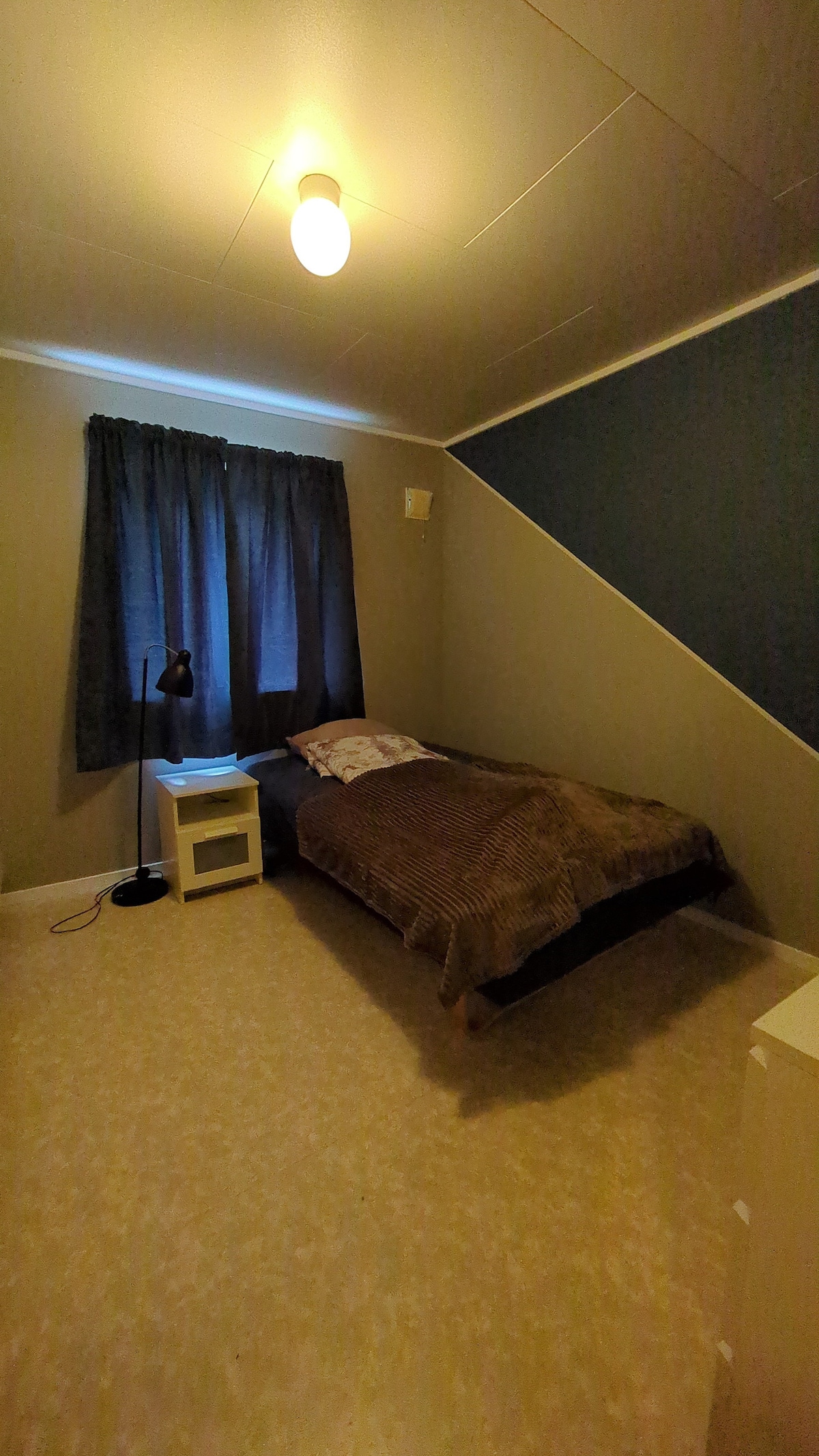 A cozy bedroom features a single bed dressed in a soft brown blanket, with a pillow at the head. A lamp stands beside a small nightstand. A curtain-covered window allows for muted light to enter, while a chair is positioned in the corner.