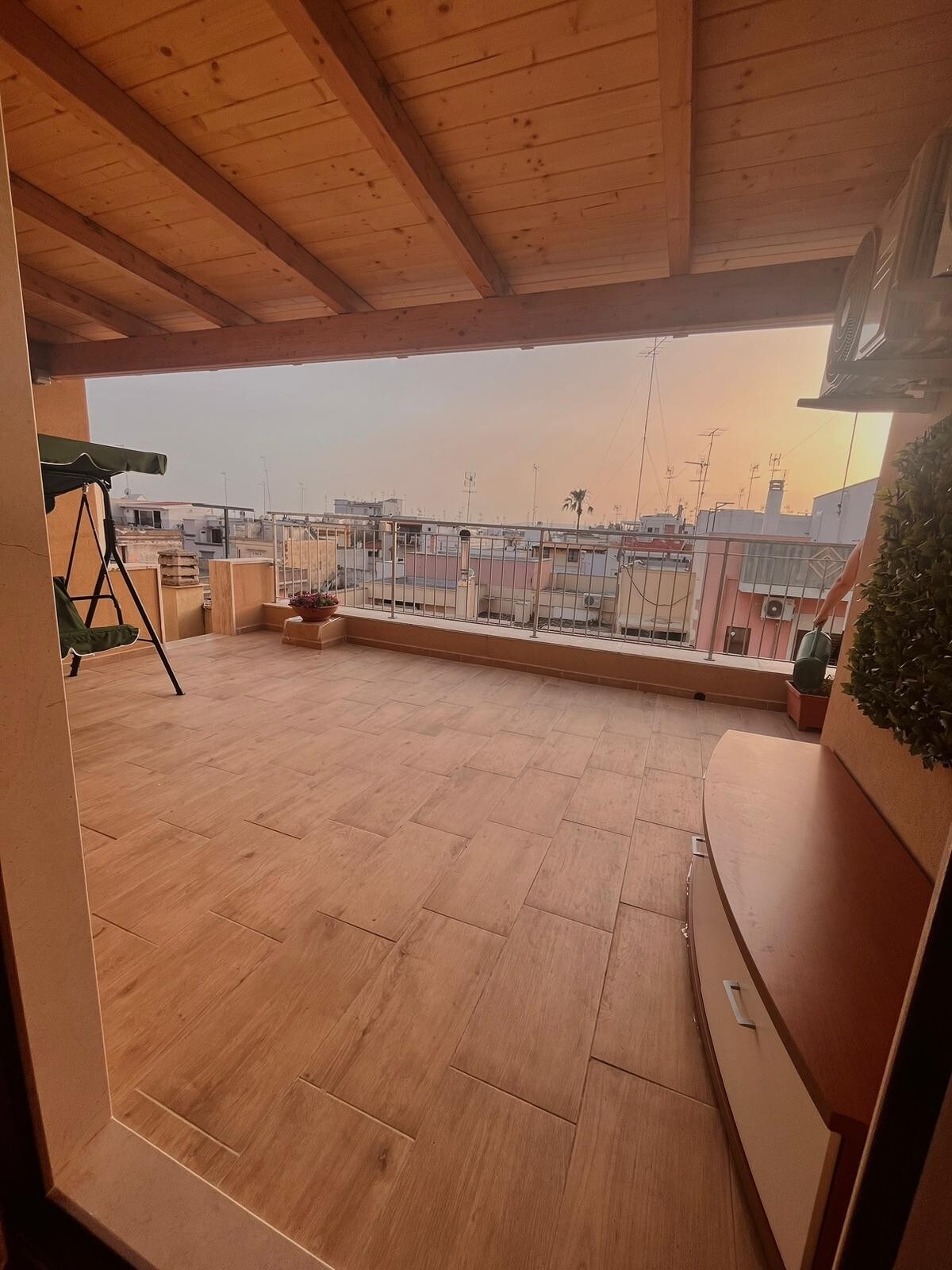 A spacious terrace features a wooden ceiling and tiled flooring, offering an expansive outdoor area. A green swing chair is positioned on one side, while planters with greenery add a touch of nature. City views are visible beyond the railing.
