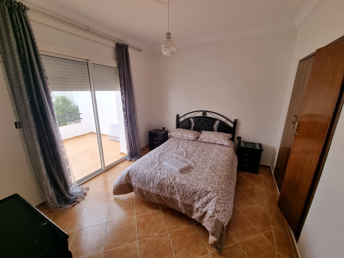 A well-appointed bedroom features a spacious bed dressed in neutral linens, complemented by bedside tables. Natural light filters through a large sliding door, which opens to a private terrace, with light curtains framing the view. The room's tiled floor contributes to a clean aesthetic.