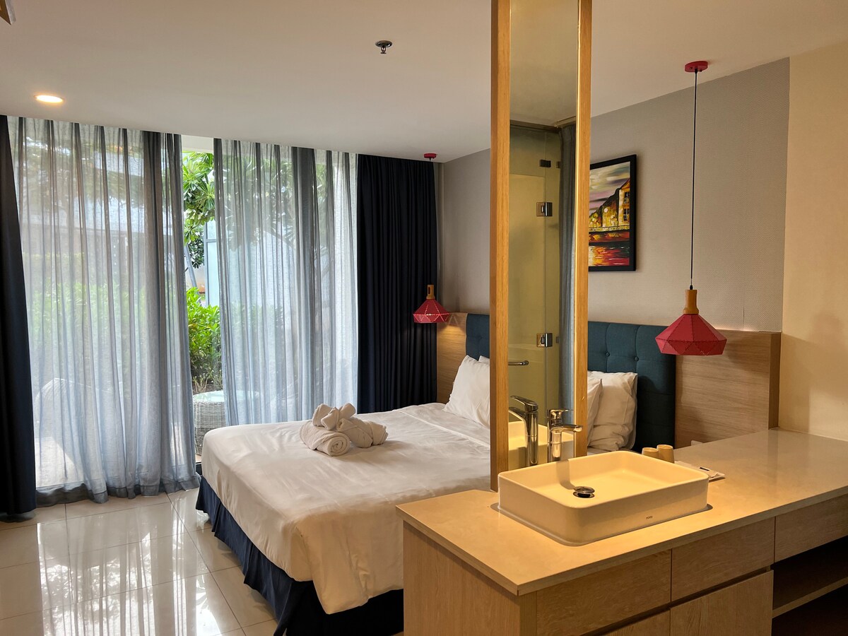 A well-appointed sleeping area features a spacious bed with neatly arranged towels, flanked by overhead pendant lights. Floor-to-ceiling windows invite natural light, revealing glimpses of greenery outside. A minimalist sink area complements the modern decor.