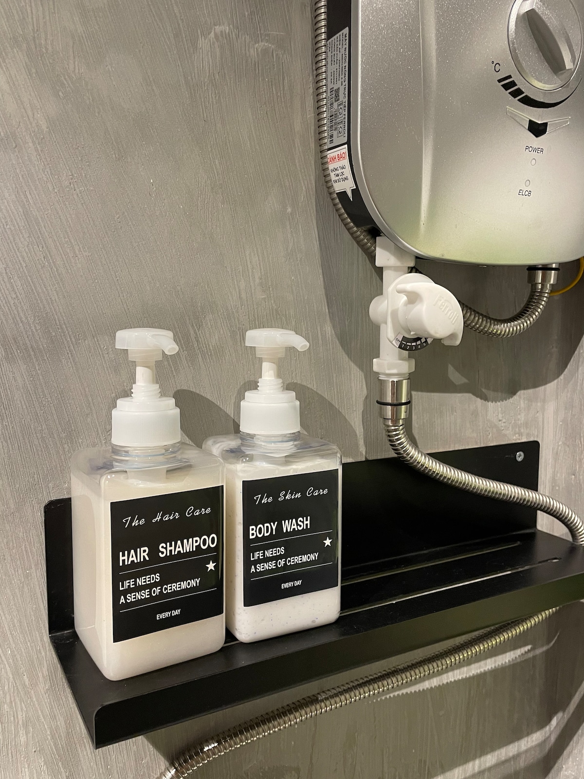 Two labeled dispensers are mounted on a dark shelf, containing hair shampoo and body wash. A chrome showerhead and an attached hose are visible above, set against a textured wall. The design provides a functional and organized bathing experience.