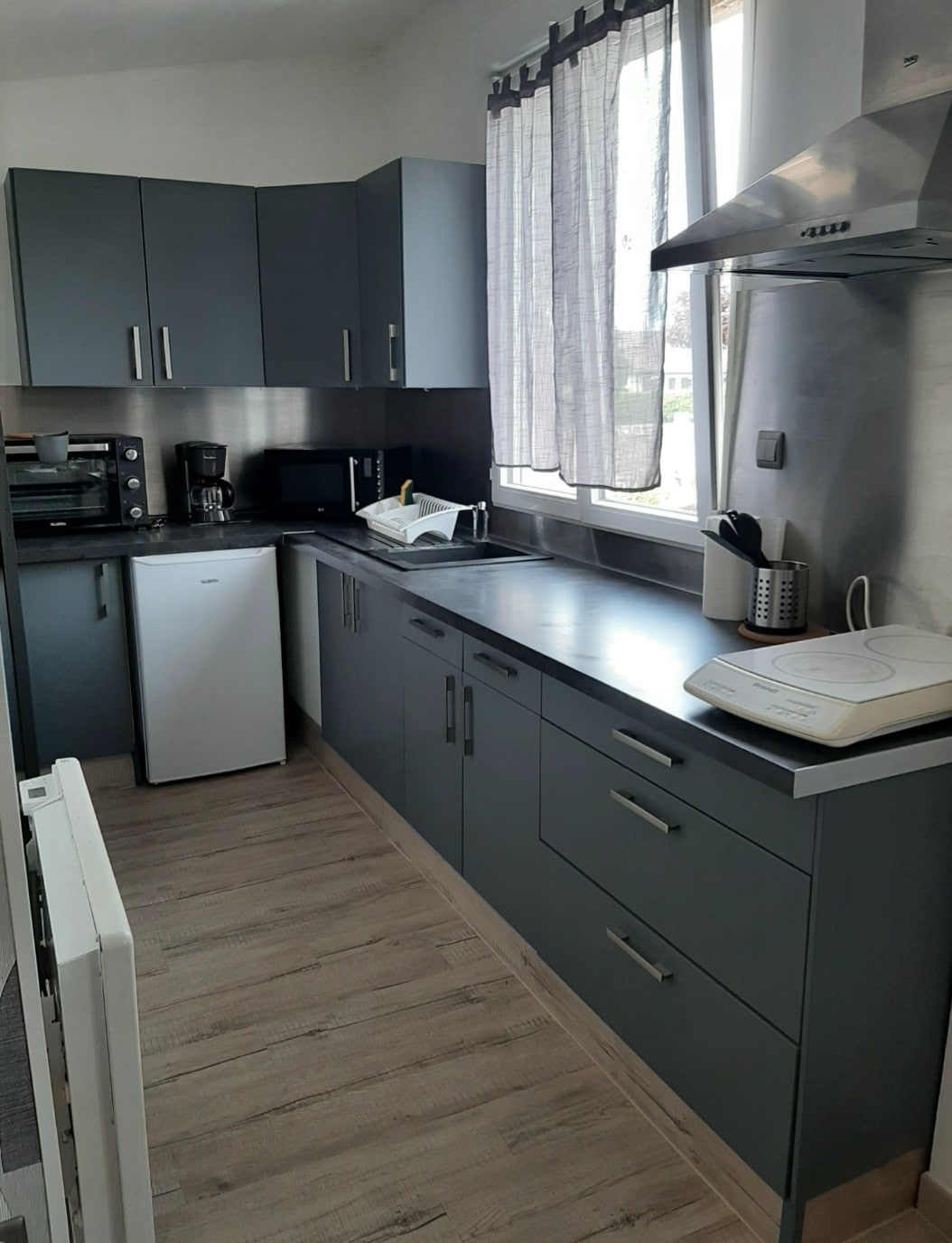 A modern kitchenette is equipped with sleek grey cabinets and a stainless steel countertop. White appliances, including a refrigerator and microwave, are visible. Natural light filters through large windows adorned with light grey curtains, illuminating the cooking space.