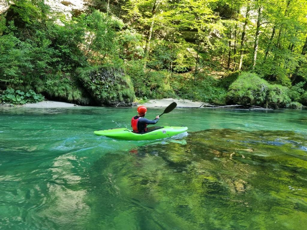 A kayaker navigates through calm, clear waters surrounded by lush greenery. Sunlight filters through the trees, creating dappled patterns on the water's surface. The vibrant green kayak contrasts with the tranquil blue and green hues of the river.