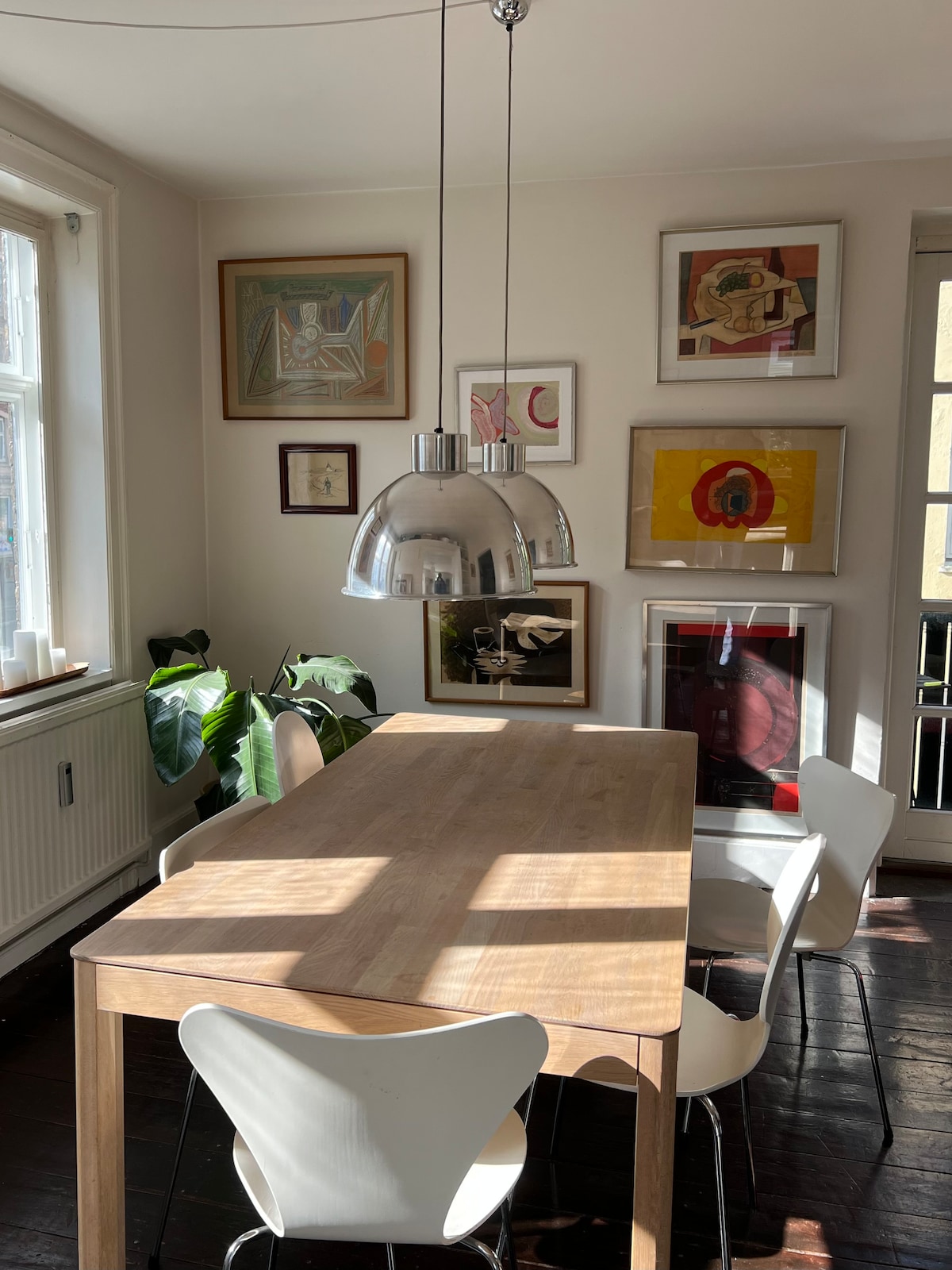 A dining area is highlighted by a wooden table surrounded by four white chairs. Two large pendant lights hang above the table, casting soft light on the surface. The walls are adorned with framed artwork, and natural light streams through the windows.