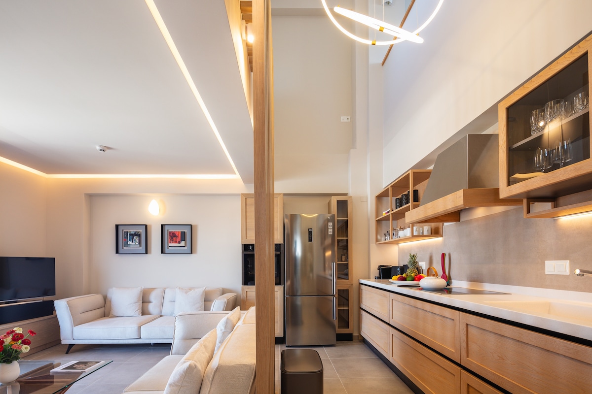 A modern living area and kitchen are showcased, featuring wooden cabinetry and sleek appliances. A comfortable sectional sofa is positioned before a television. Natural light is allowed in through large windows, creating an inviting and open space that connects the living room to the kitchen.