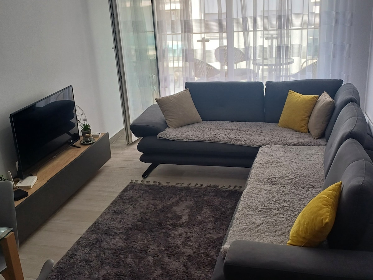 A bright living area features a spacious sectional sofa adorned with yellow cushions and a soft, textured throw. A sleek media console holds a television, while natural light filters through sheer curtains, revealing a balcony space with outdoor seating.