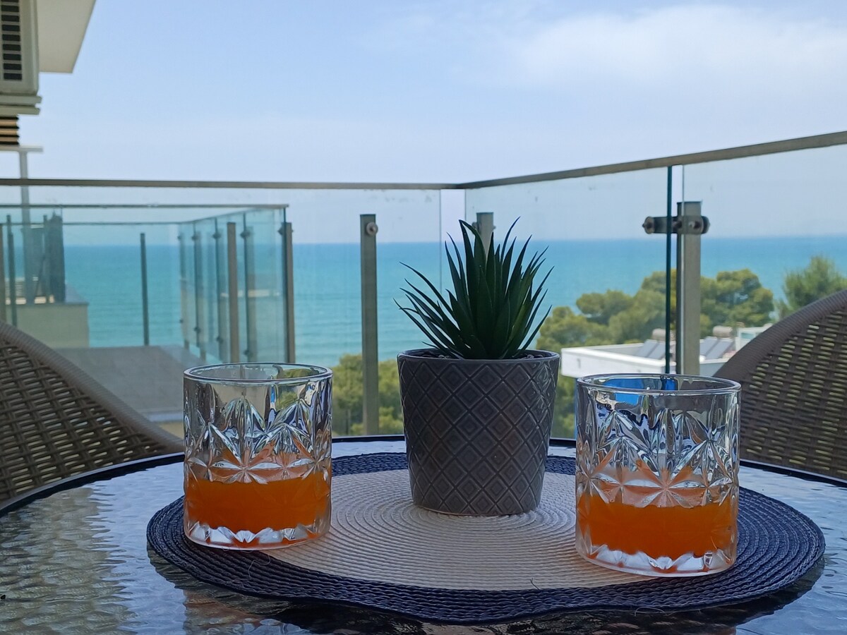 A glass table is set with two crystal tumblers filled with orange beverage and a small potted plant at the center. In the background, the serene blue sea and green treetops are visible, framed by a glass balcony railing.